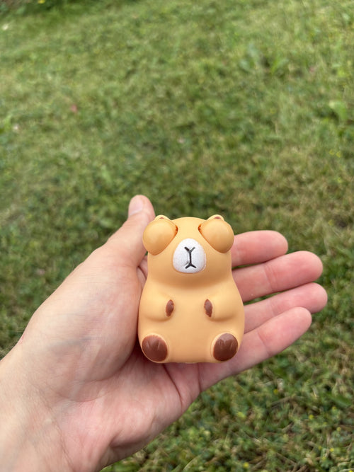 A small capybara squishy held in a person's hand against a blurred green grass background.