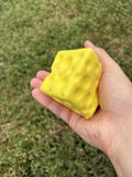 A hand holding a bright yellow, cheese-squishy toy against a blurred green grass background.